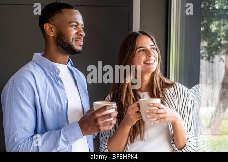 Una coppia sorridente in piedi vicino a una finestra con tazze di caffè, godendosi una mattinata rilassata insieme a casa, condividendo una conversazione calda e uno sguardo speranzoso Foto Stock