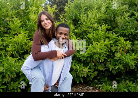 Giovane coppia che ride e si gode un gioioso giro a cavalluccio in un ambiente esterno lussureggiante. Abiti casual, sorrisi caldi e linguaggio del corpo affettuoso con Foto Stock
