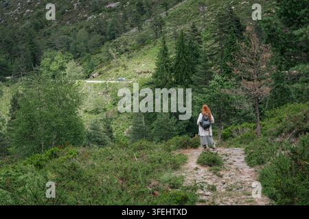 Fotografia esterna di vista posteriore di un'escursionista con lunghi capelli rossi che porta uno zaino mentre cammina lungo un sentiero roccioso su un pendio montano lussureggiante. Eva Foto Stock