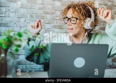 Donna dai capelli ricci con cuffie e occhiali gode della musica mentre lavora su un laptop in un accogliente ufficio domestico, mostrando una produttività rilassata, concentrazione, a Foto Stock