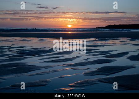 Fotografia di paesaggio costiero ad angolazione bassa che cattura un vivido tramonto dorato sull'orizzonte dell'oceano. I raggi del sole si riflettono nelle pozze di marea poco profonde e Ripp Foto Stock