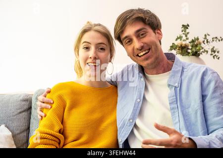 Giovane coppia felice seduta insieme su un divano a casa, sorridente e abbracciata in un ritratto casual e accogliente Foto Stock