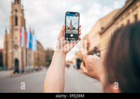 Novi Sad, Serbia – 23 luglio 2026: Turista femminile a Novi Sad che scatta foto con lo smartphone del punto di riferimento, la Chiesa del Nome di Maria. Foto Stock