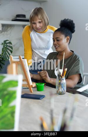 Diverse amiche che dipingono su piccole tele nello studio di casa con cavalletto da tavolo e barattolo per pennelli Foto Stock