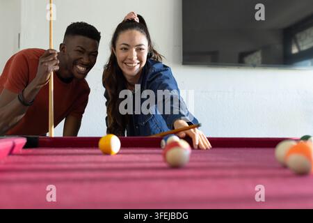 Donna asiatica e uomo afroamericano che giocano a biliardo in una sala ricreativa a un tavolo magenta tenendo le stecche. Biliardo, duo, casual, amicizia, competizione, l Foto Stock