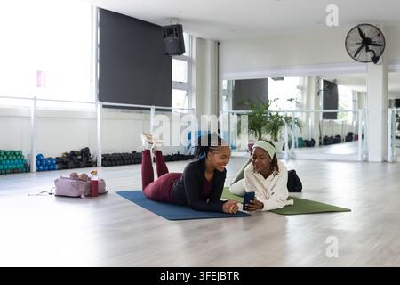 Due donne africane sdraiate su materassini da yoga in una palestra, indossano leggings bordeaux, controllano il telefono Foto Stock