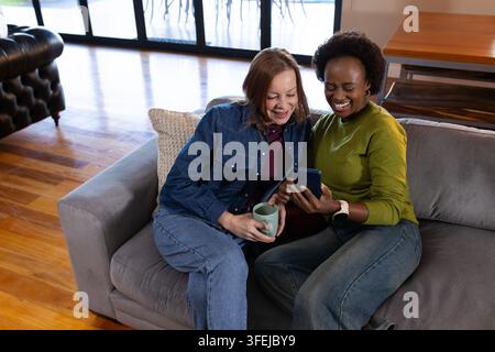 Diverse amiche femminili sedute su un divano grigio, appoggiate e sorridenti mentre guardano il telefono, tenendo una tazza Foto Stock