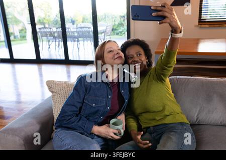 Diverse amiche femminili sedute vicine su un divano grigio che scattano un selfie con uno smartphone e una tazza Foto Stock