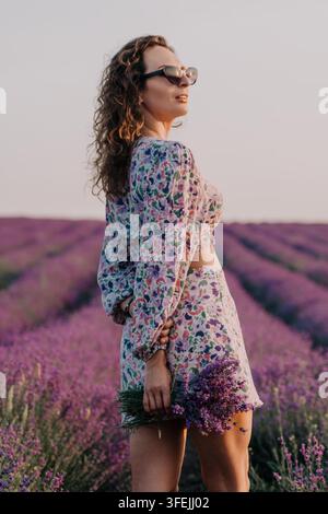 Una giovane donna in un set floreale a due pezzi e occhiali da sole si trova in un vivace campo di lavanda viola al tramonto, tenendo un mazzo di steli di lavanda. Foto Stock