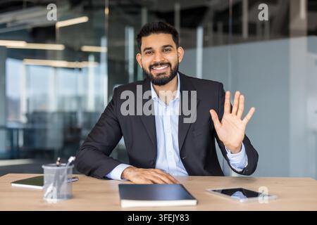 Uomo d'affari indiano seduto a una scrivania moderna in ufficio, sorride alla telecamera e fa un gesto di saluto, accoglie clienti o colleghi durante una videoconferenza online Foto Stock