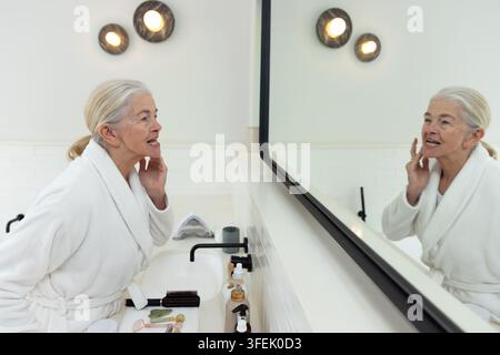 Donna anziana in piedi al lavandino del bagno, tocca la guancia mentre guarda nello specchio con strumenti per il viso Foto Stock