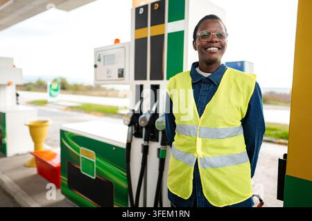 Addetto alla stazione di servizio con indosso un giubbotto di sicurezza e sorridente accanto alle pompe di benzina Foto Stock