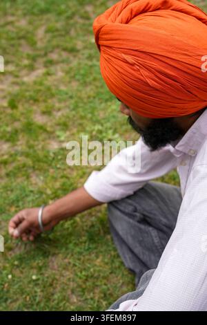 Prima piano ad angolazione alta di un giovane uomo sikh con un turbante arancione vivace, seduto su un'erba verde rigogliosa e raccogliendo con cura un piccolo fiore selvatico. Foto Stock