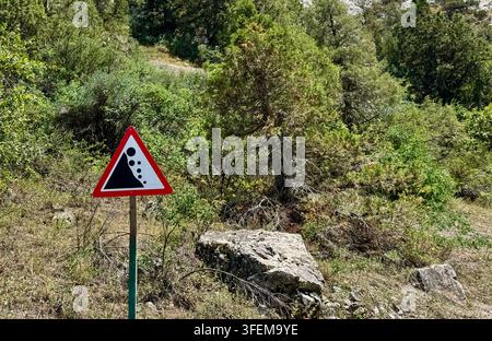 Segnale triangolare di avvertimento per caduta massi si trova accanto a un pendio boscoso ripido con massi sparsi, erba secca e arbusti illuminati dal sole, illustra la strada Foto Stock