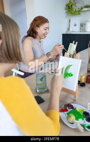 Amiche che ridono, dipingendo su piccoli cavalletti da tavolo al tavolo della cucina con tavolozza Foto Stock