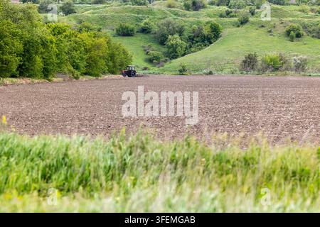 Trattore agricolo che lavora in un campo marrone coltivato accanto ad alberi verdi rigogliosi e dolci colline erbose sotto un cielo diurno luminoso con sfocato per Foto Stock