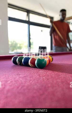 Uomo africano con maglietta arrugginita che tiene una stecca in una sala da gioco vicino a palle disposte su un feltro bordeaux Foto Stock