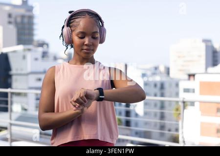 Donna adulta africana in piedi su un tetto vicino a una rotaia che controlla un orologio nero indossando un auricolare rosa e un crop top Foto Stock