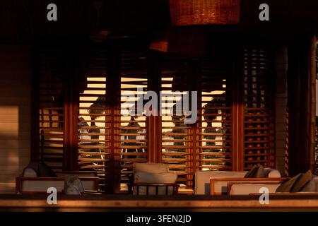 La luce calda del tramonto riempie un ristorante di legno all'aperto con tavoli vuoti, sedie intrecciate, sgabelli da bar, lampade sospese e vegetazione tropicale. Foto Stock
