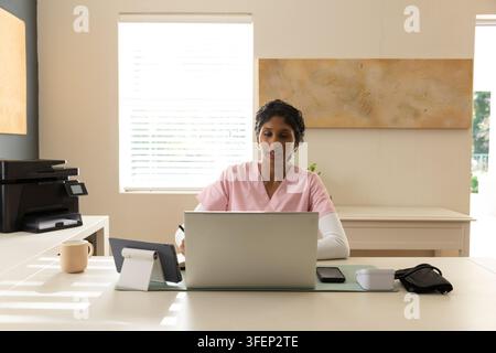 Donna indiana in camici rosa che lavora su un laptop alla scrivania con una tazza tablet e uno smartphone Foto Stock