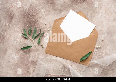 Modello di invito per matrimonio. Biglietto bianco vuoto in una busta kraft, nastro, ramo d'olivo e perle. Sfondo beige grunge. Vista dall'alto, flat lay Foto Stock