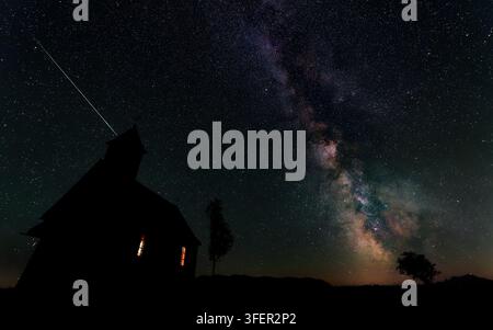 Una meteora delle Perseidi si staglia nel cielo notturno sopra la cappella illuminata di Gahberg, con la Via Lattea che sorge sulla regione di Attersee, in Austria Foto Stock