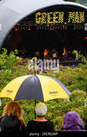GUARDARE IL PALCO SOTTO LA PIOGGIA AL FESTIVAL GREEN MAN, REPORTAGE, FESTIVAL GREEN MAN 2012, BRECON, GALLES, REGNO UNITO, 2012: Di spalle, diversi partecipanti al festival guardano una band esibirsi sul palco principale illuminato durante un acquazzone, uno tiene alto un ombrello giallo e nero. Foto: Rob Watkins. INFO: Il festival Green Man si tiene annualmente ad agosto nelle Brecon Beacons. Celebra la musica alternativa, le arti, la letteratura e la scienza in un ambiente montano adatto alle famiglie. Il festival del 2012 è stato il più fangoso mai registrato dopo una settimana di forti piogge. Foto Stock