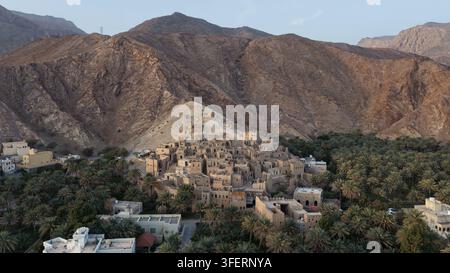 Vista aerea delle rovine storiche in mattoni di fango di Birkat Al Mouz incastonate in un'oasi lussureggiante di palme, sullo sfondo di montagne aspre a Birkat A Foto Stock