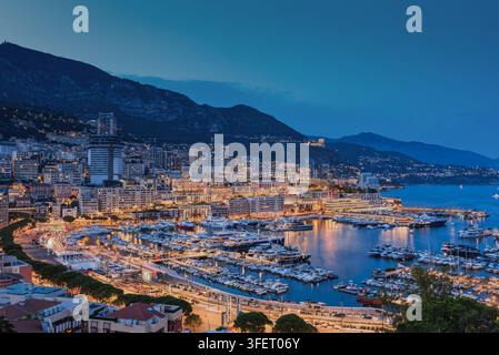 Vista aerea del Port Hercules a Monaco al crepuscolo, con yacht di lusso ormeggiati nel porto, edifici cittadini illuminati e montagne sullo sfondo Foto Stock