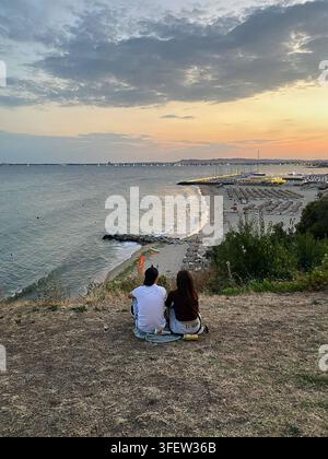 Saint Vlas, Bulgaria – 13 agosto 2026: Coppia seduta su una pendice erbosa che osserva il tramonto sulla spiaggia con sedie a sdraio e onde dell'oceano in basso Foto Stock