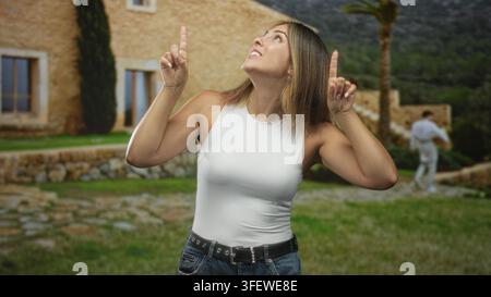 La donna punta le dita indice verso l'alto con le mani visibili e sorride di fronte all'edificio in pietra, indossando canotta bianca e jeans; gioia giocosa. Foto Stock