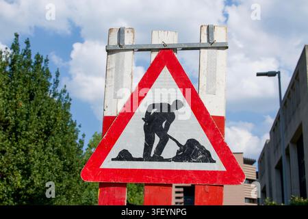 Segnale di pericolo di lavori stradali davanti con un pittogramma di un uomo che scava montato su una barriera del cantiere contro un cielo nuvoloso. Concetto per la costruzione stradale Foto Stock