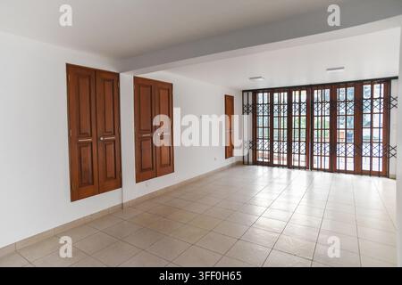 Stanza vuota con porte e finestre in legno: Uno spazio interno non arredato con pareti bianche, pavimento piastrellato e grandi porte in legno con persiane Foto Stock