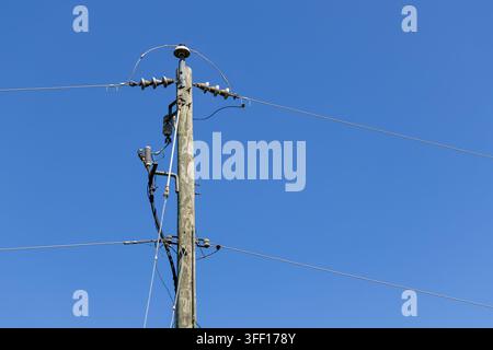 Vista ravvicinata di un palo elettrico in legno con isolatori, apparecchiature elettriche e linee di distribuzione aeree silouettate contro un cielo blu chiaro Foto Stock