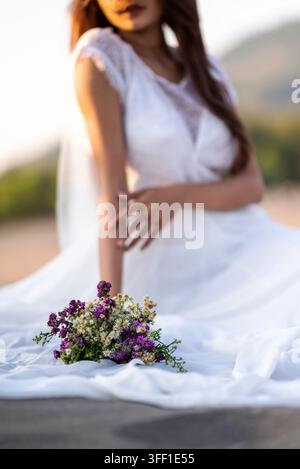 Primo piano di un bouquet nuziale rustico di fiori selvatici viola e bianchi, appoggiato sul tessuto di un vestito bianco con una sposa sfocata sullo sfondo dur Foto Stock