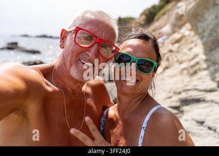Coppia di anziani felici con occhiali da sole che posano per un selfie in vacanza estiva al mare Foto Stock