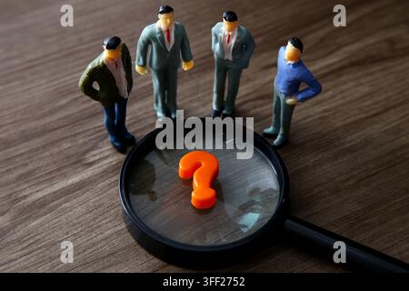 Piccole persone d'affari in miniatura che collaborano per risolvere un problema, concentrandosi su un punto interrogativo arancione con una lente d'ingrandimento Foto Stock