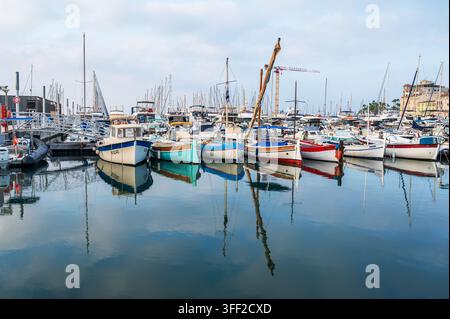 Cannes, Francia - 2 agosto 2026: Barche da pesca tradizionali in legno e yacht moderni ormeggiati in un porto tranquillo con riflessi sull'acqua. Foto Stock