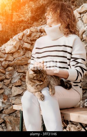 Donna che accarezza una giovane donna in maglione a righe siede su una panchina con un felino tigrato alla luce dorata del sole vicino a un muro di pietra Foto Stock