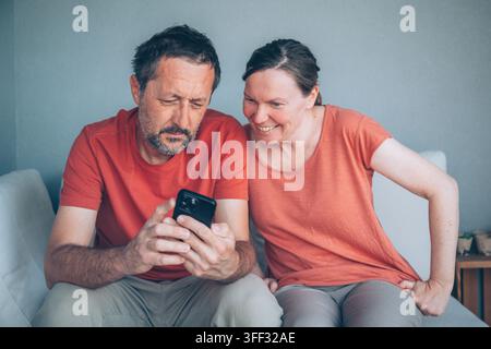 Coppia felice di mezza età che guarda insieme lo smartphone sul divano, condividendo contenuti online divertenti e godendosi il tempo a casa, messa a fuoco selettiva Foto Stock