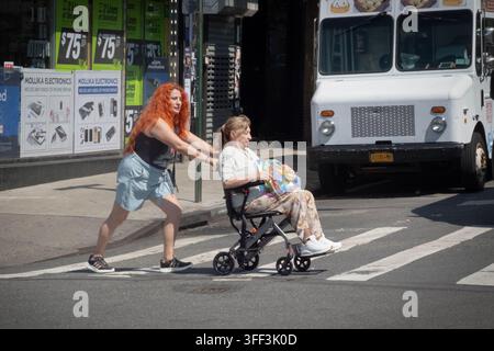 Una donna con lunghi capelli rossi spinge un'anziana donna seduta su un deambulatore attraverso la 74th Street a Jackson Heights, Queens, New York. Foto Stock