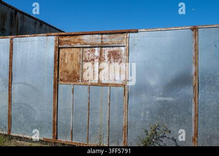 un vecchio muro di metallo consumato costruito con pannelli in acciaio zincato incorniciati da travi in ferro pesantemente arrugginite sotto un cielo blu luminoso e sereno. Foto Stock