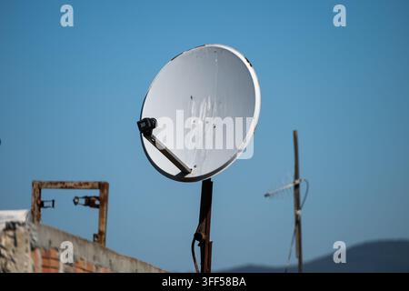 Una parabola satellitare bianca consumata montata su un palo arrugginito su un tetto contro un cielo blu chiaro, con un'antenna televisiva sfocata visibile sullo sfondo Foto Stock