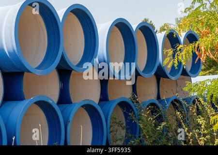 Una pila di grandi tubi industriali blu con interni bianchi si trova all'aperto, parzialmente oscurata da fogliame verde e giallo contro un cielo luminoso. Foto Stock