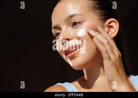 Una giovane donna sorride mentre applica la crema per il viso, evidenziando una routine di cura della pelle per una pelle sana e radiosa. Foto Stock