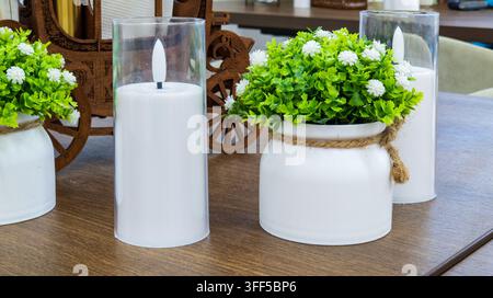 Decorazione da tavolo in un caffè. Candele bianche in cilindri di vetro sono posate su un tavolo di legno accanto a piante artificiali in vaso. Arredamento interno accogliente di un ristorante Foto Stock
