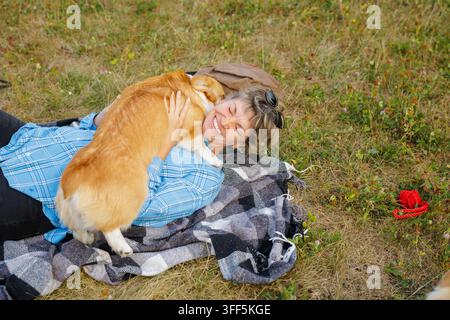 Donna anziana sorridente sdraiata su una coperta all'aperto mentre abbraccia il suo affettuoso cane corgi, illustrando la compagnia, la terapia con gli animali e l'invecchiamento attivo le Foto Stock