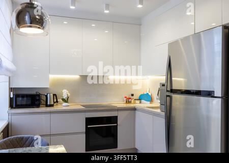 Vista interna di una cucina moderna e ben illuminata con mobili bianchi lucidi e illuminazione calda sotto i mobili. Il design contemporaneo include una b Foto Stock