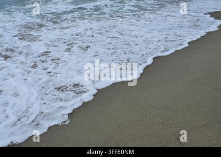 Primo piano di acqua marina schiumosa che rotola sulla linea di riva di una spiaggia sabbiosa bagnata Foto Stock