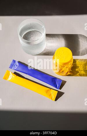 Bicchiere d'acqua con pillole di integrazione in barattolo e bustine di polvere vitaminica su tavolo beige alla luce del sole. Concetto di alimentazione sana e farmacia. Vitamina la Foto Stock